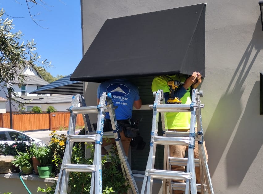 Two workers focused on installing a black awning on a house, enhancing the home's exterior with a functional feature.